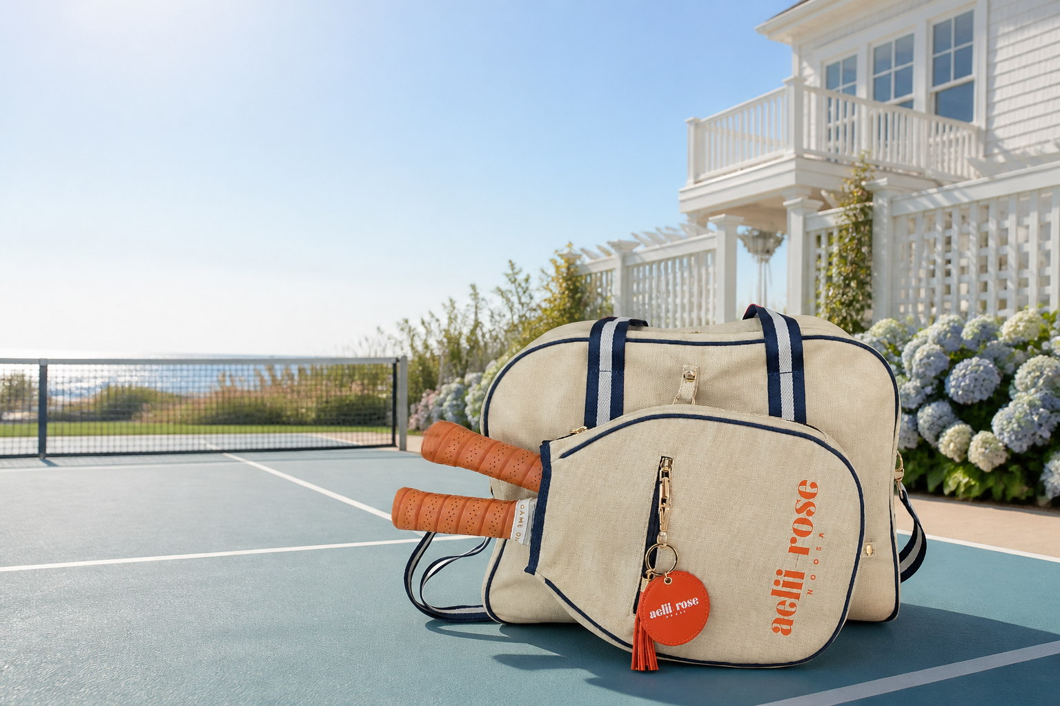 Aelii-Rose Noosa pickleball totes and paddle sleeve on a coastal court with a Hamptons-style house in the background